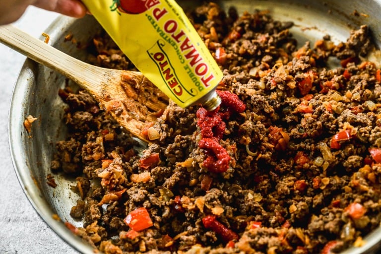 A hand squeezes tomato paste from a tube into a skillet of cooked ground beef and vegetables being stirred with a wooden spoon.