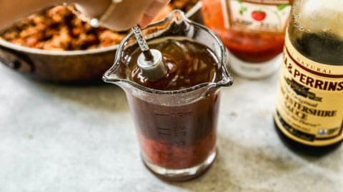 Hand stirring a sloppy joe sauce sauce in a glass measuring cup, with ketchup and Worcestershire sauce bottles nearby.