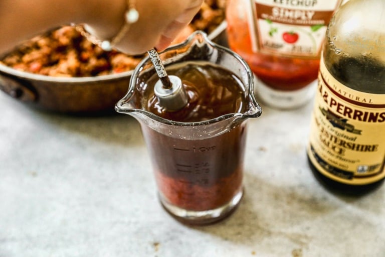 Hand stirring a sloppy joe sauce sauce in a glass measuring cup, with ketchup and Worcestershire sauce bottles nearby.