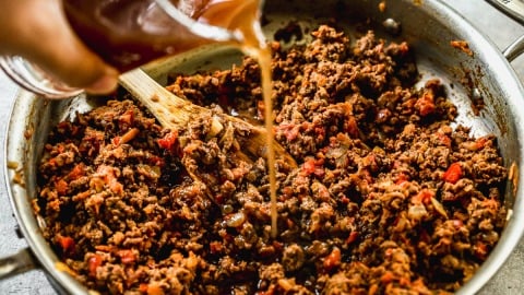 A hand pours broth into a skillet with cooked ground beef, onions, and red bell peppers, stirring with a wooden spoon.