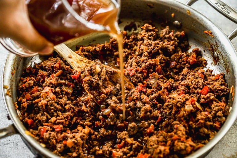 A hand pours broth into a skillet with cooked ground beef, onions, and red bell peppers, stirring with a wooden spoon.