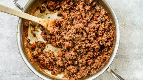 Cooked ground beef sloppy joe filling with tomato sauce being stirred by a wooden spoon in a stainless steel skillet.