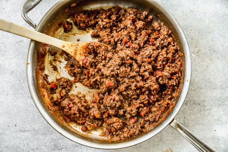 Cooked ground beef sloppy joe filling with tomato sauce being stirred by a wooden spoon in a stainless steel skillet.