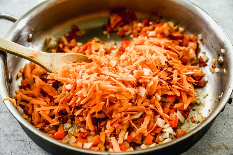 A pan with chopped onions, red bell peppers, and shredded carrots being stirred with a wooden spoon.