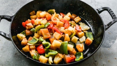 Colorful sautéed vegetables and pineapple chunks in a black skillet on a gray surface.