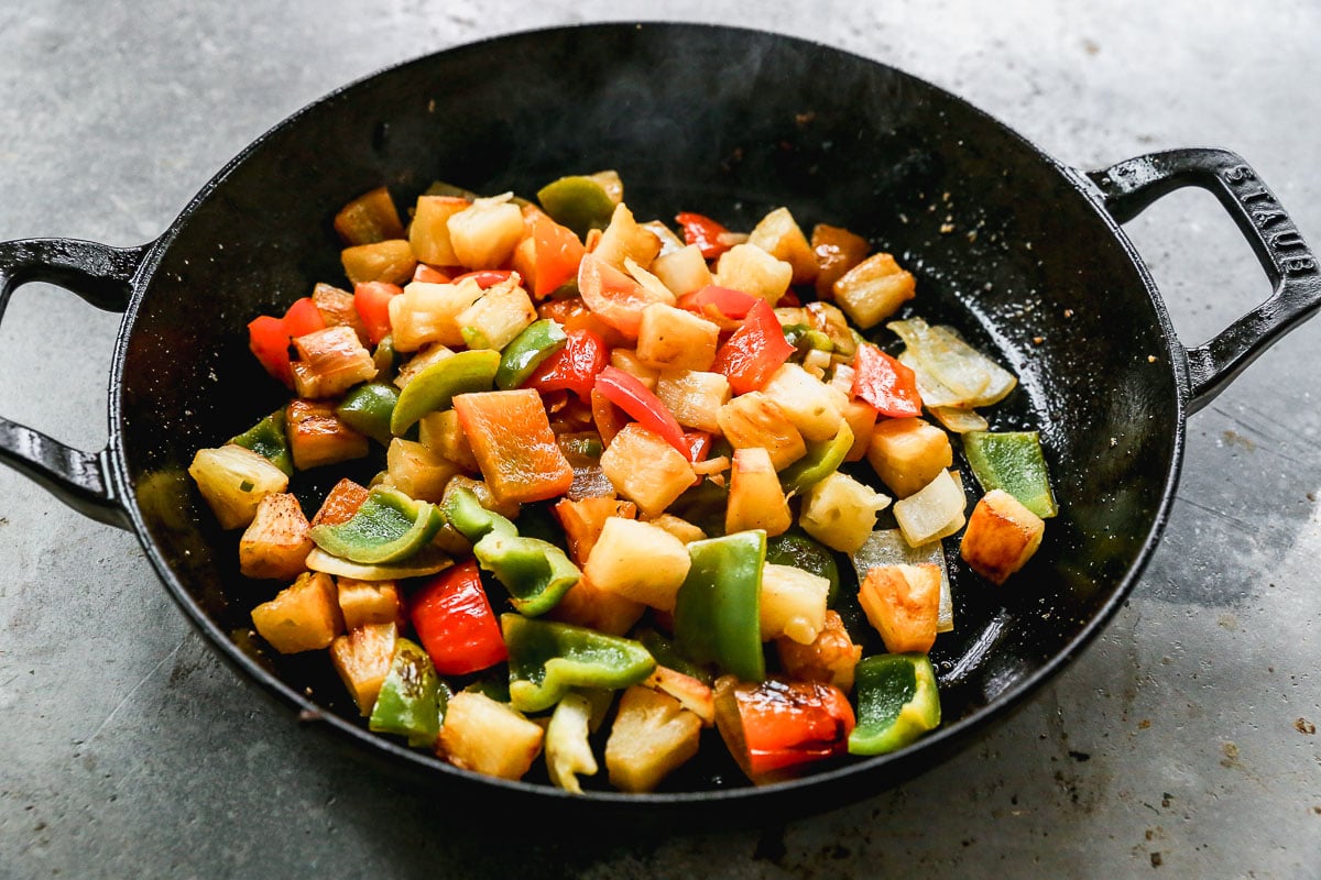 Colorful sautéed vegetables and pineapple chunks in a black skillet on a gray surface.