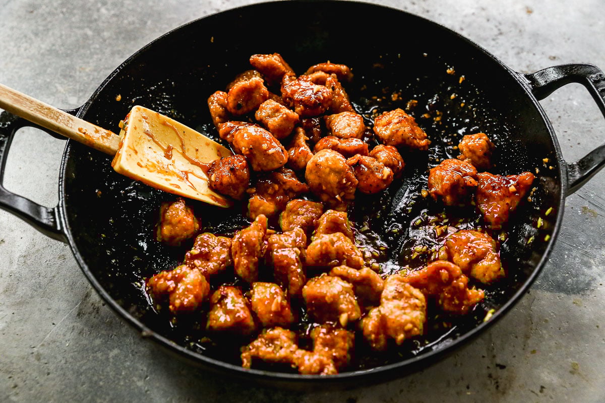Crispy pork pieces coated in sauce being stirred in a black skillet with a spatula.