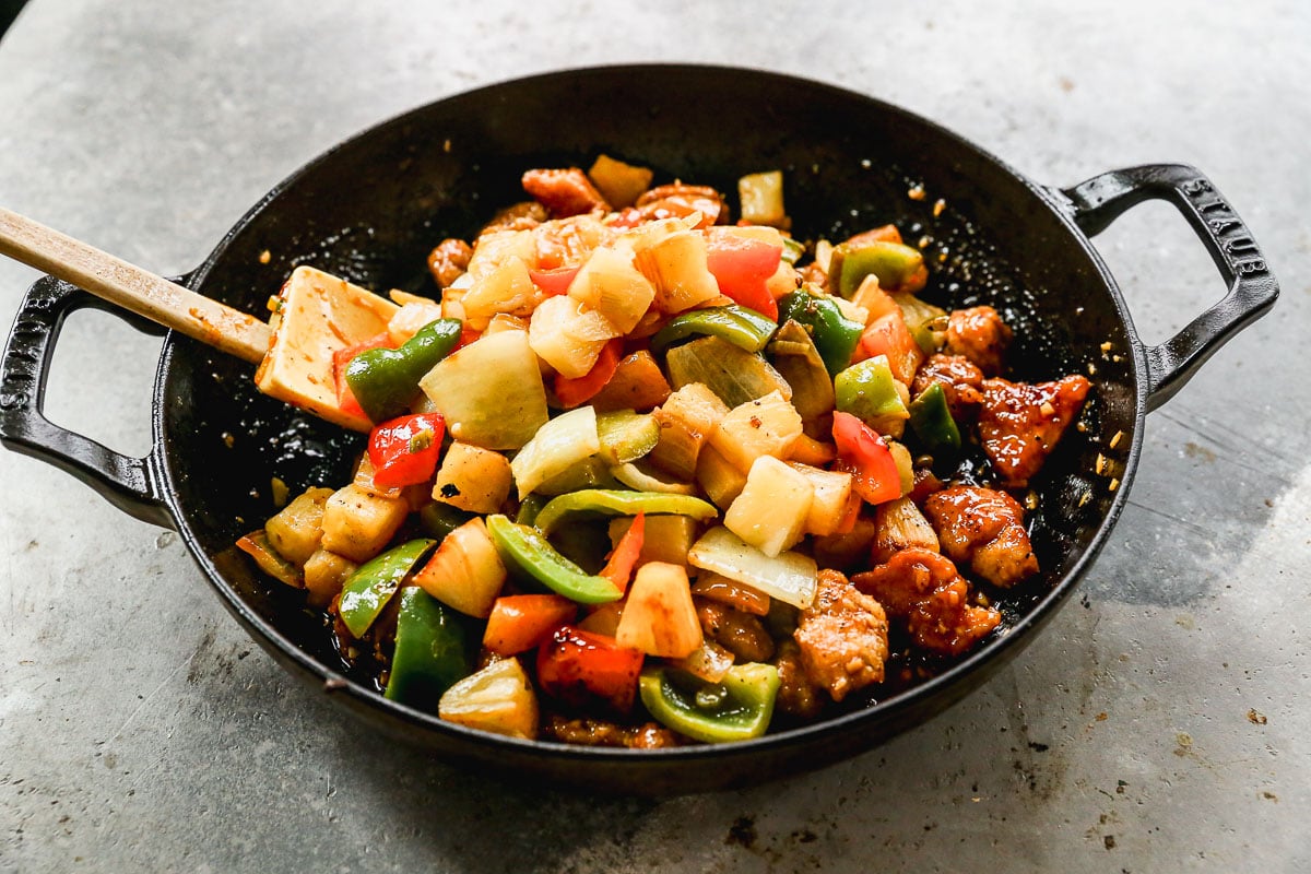 A skillet filled with sweet and sour pork, bell peppers, onions, and pineapple chunks, being stirred with a spatula.