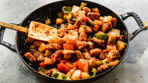 A skillet filled with sweet and sour pork, mixed with bell peppers, pineapple, and sauce, being stirred with a spatula.