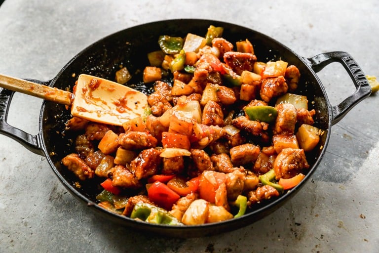 A skillet filled with sweet and sour pork, mixed with bell peppers, pineapple, and sauce, being stirred with a spatula.