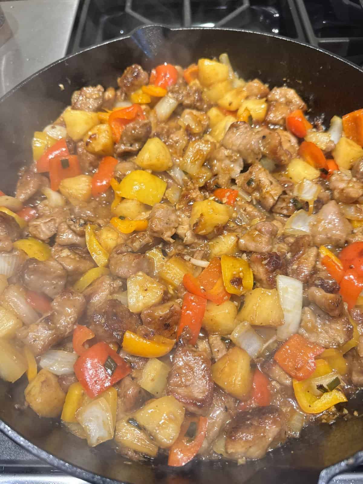 A skillet filled with cooked pork, bell peppers, onions, and pineapple chunks on a stovetop.