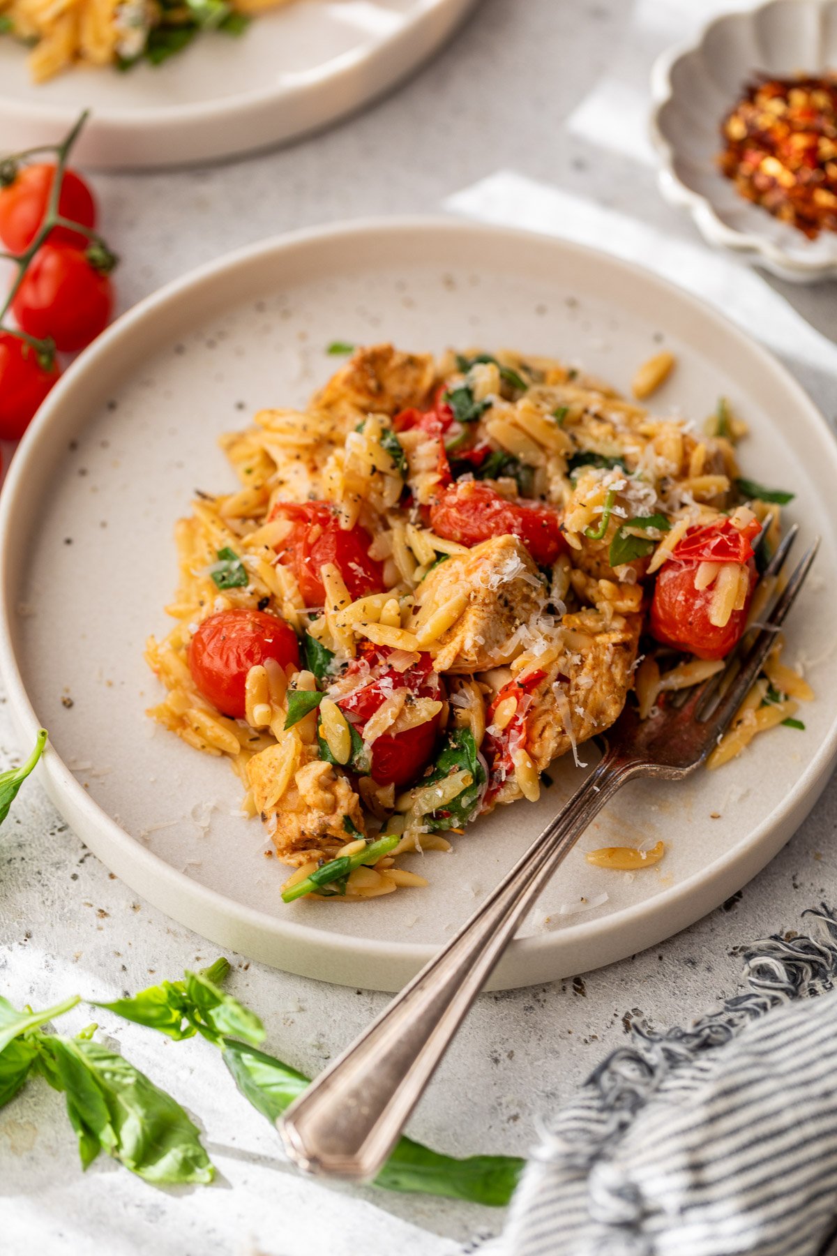 A plate of orzo with chicken, cherry tomatoes, spinach, parmesan, and herbs, served with a fork on a light-colored dish.