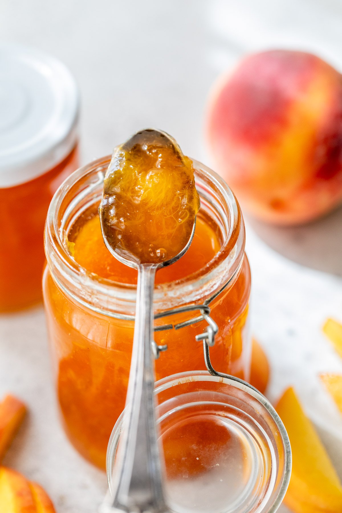 A spoonful of peach jam held over an open glass jar, with a fresh peach and jar lid in the background.