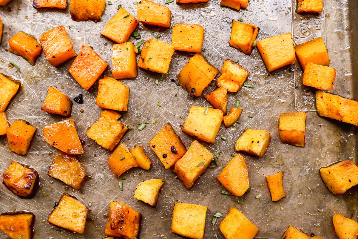 Roasted butternut squash cubes scattered on a baking sheet, seasoned with herbs and spices.