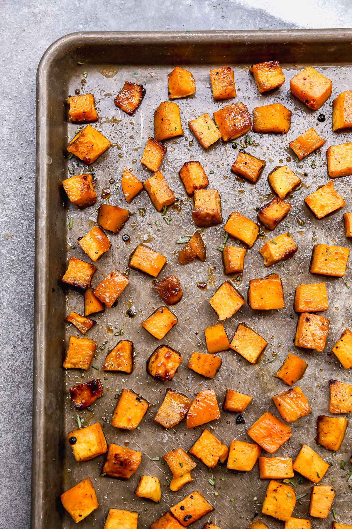 Cubed roasted butternut squash scattered on a metal baking sheet, lightly browned and garnished with herbs.