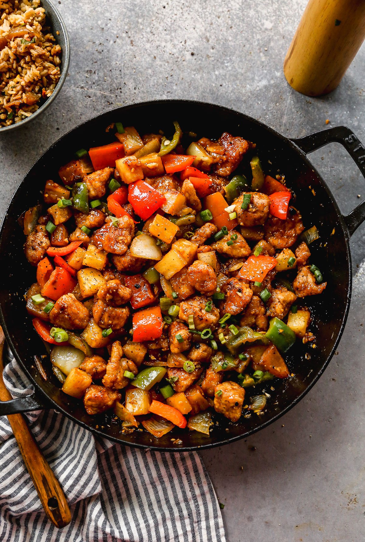 A skillet filled with stir-fried sweet and sour pork, bell peppers, pineapple, and green onions on a gray surface.
