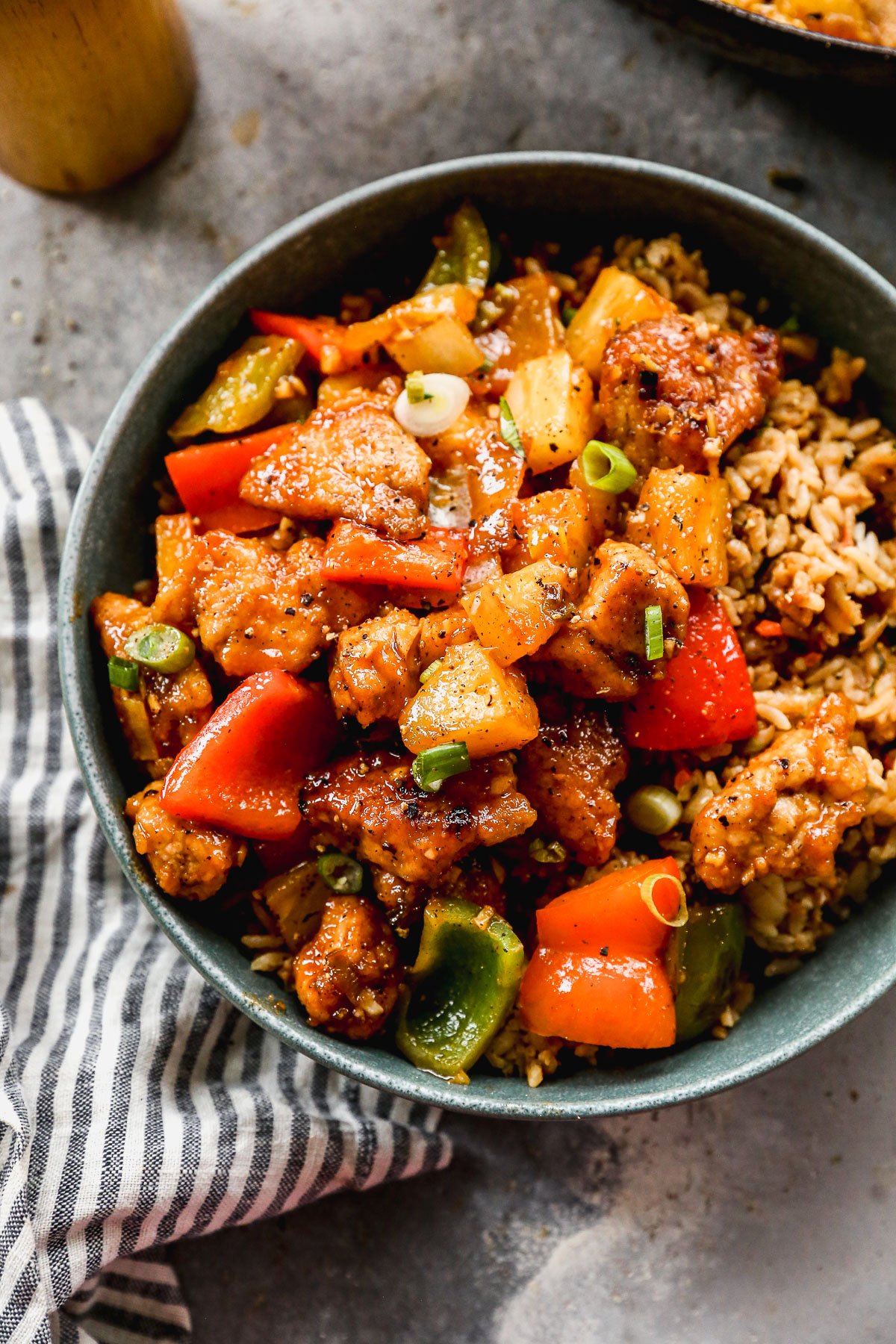 A bowl of sweet and sour pork with bell peppers, pineapple, and rice, placed on a striped napkin.
