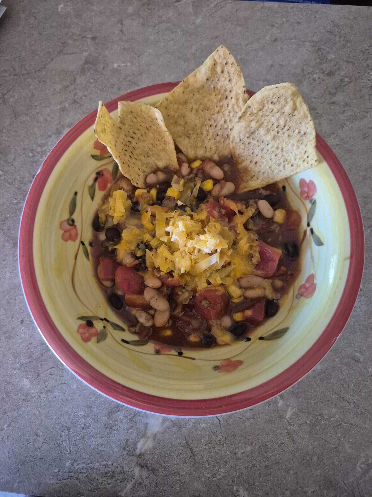 A bowl of chili with beans, corn, cheese, and tomatoes, topped with tortilla chips on a floral-patterned plate.