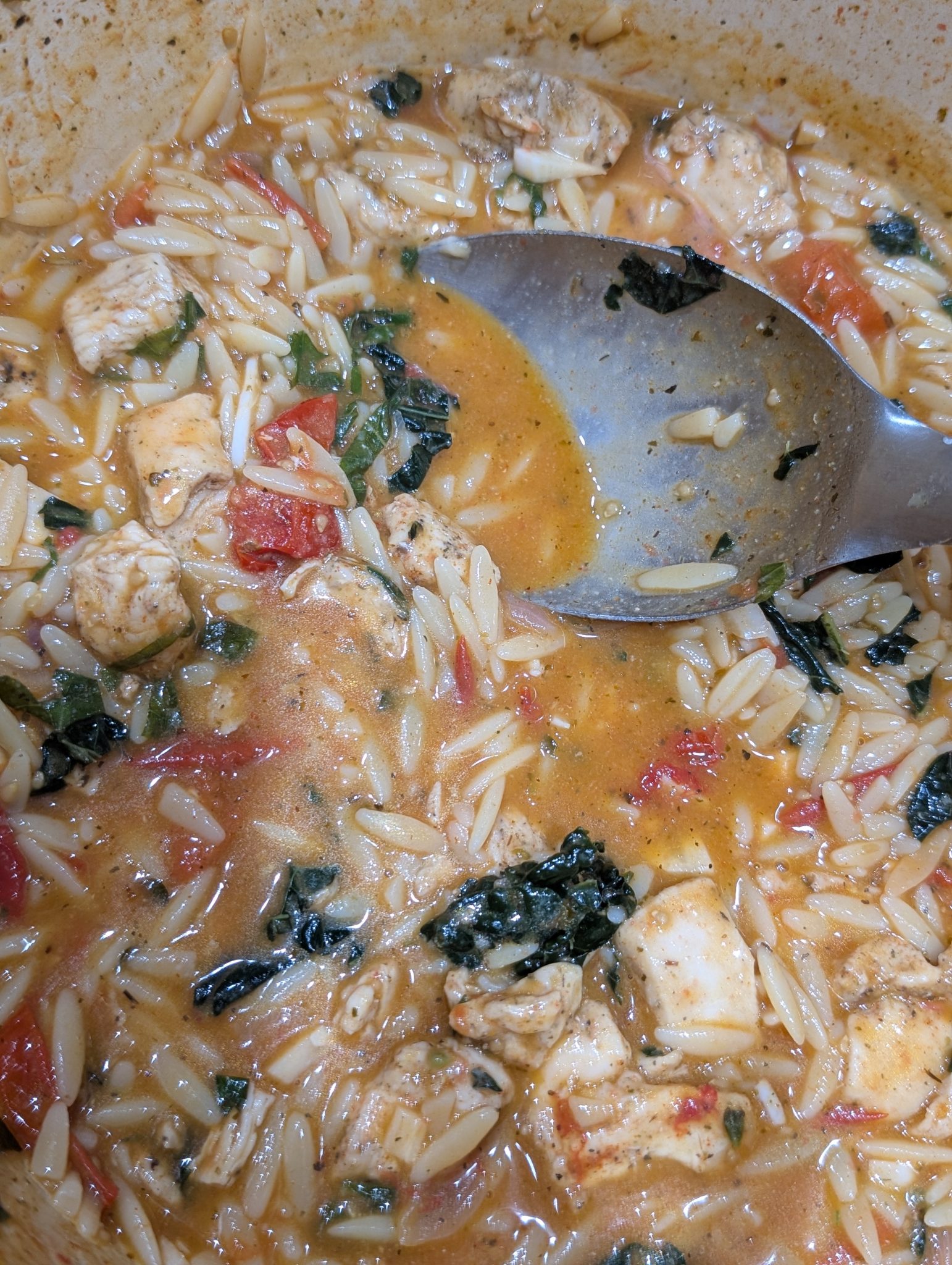 A close-up of a pot with orzo pasta, chicken, tomatoes, leafy greens, and a metal spoon in a creamy broth.