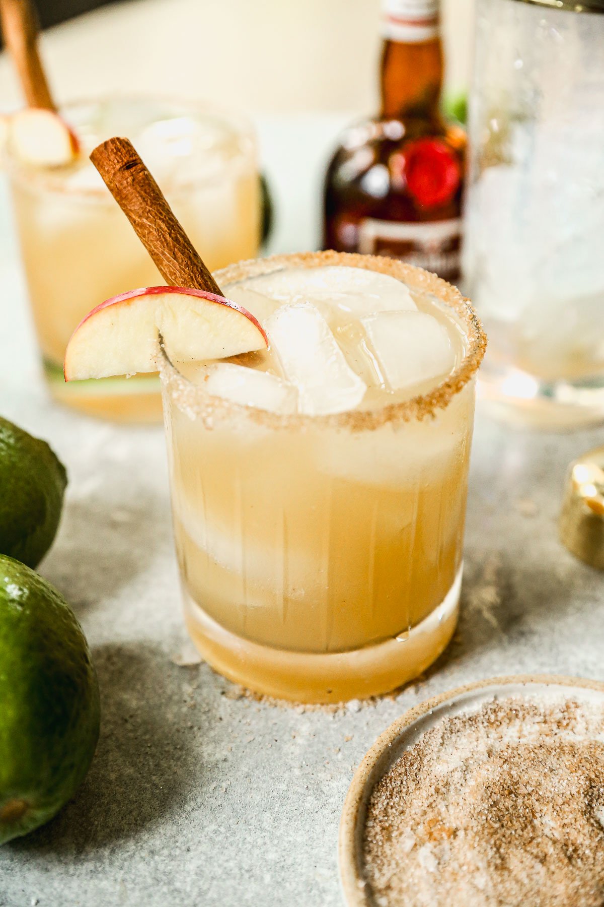 An apple cider margarita with ice, a cinnamon stick, and apple slice in a glass with a spiced rim; limes and a liquor bottle nearby.