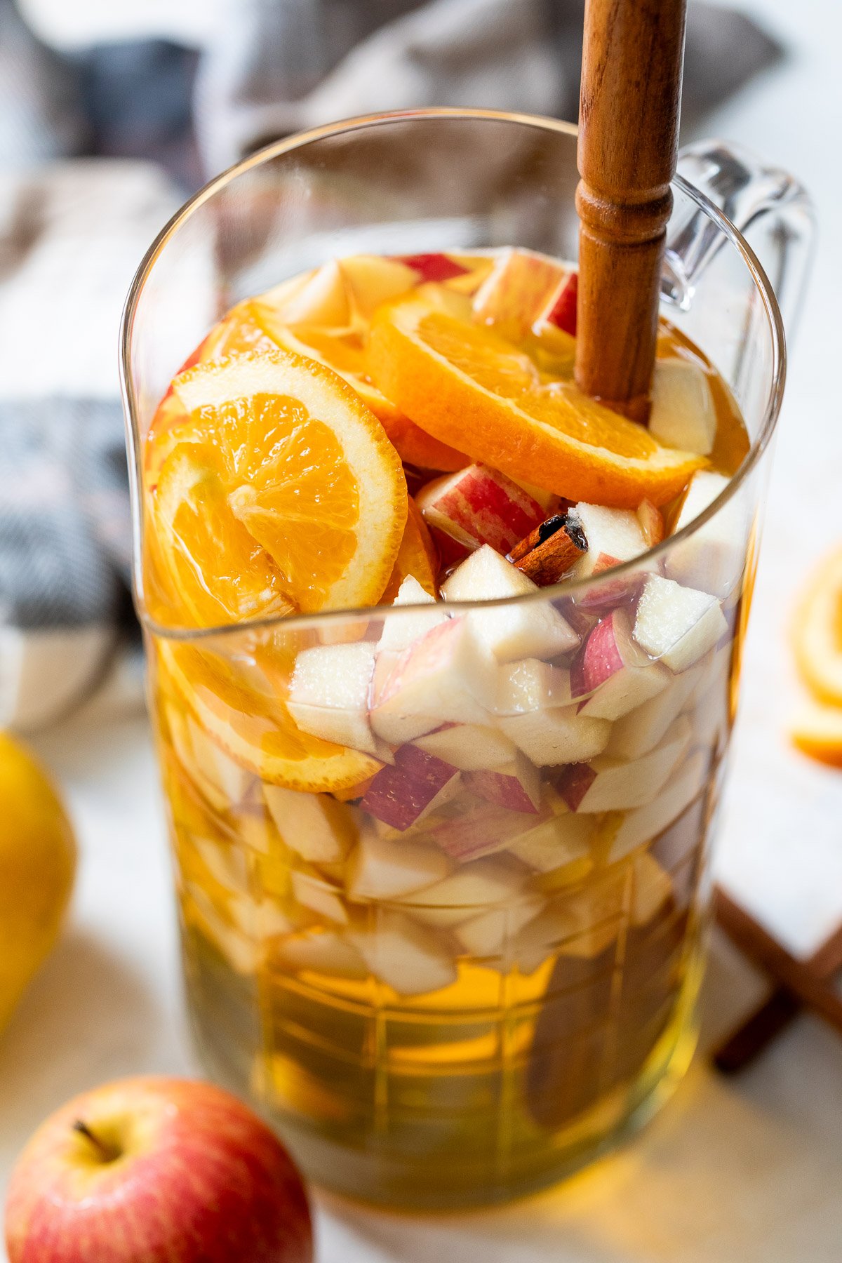 A glass pitcher filled with apple cider sangria, sliced oranges, chopped apples, and a wooden spoon for stirring.