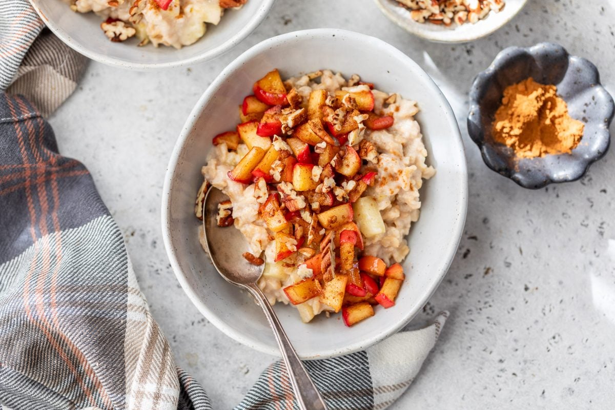 A bowl of apple cinnamon oatmeal topped with diced apples, pecans, and cinnamon, with a spoon and plaid napkin beside it.