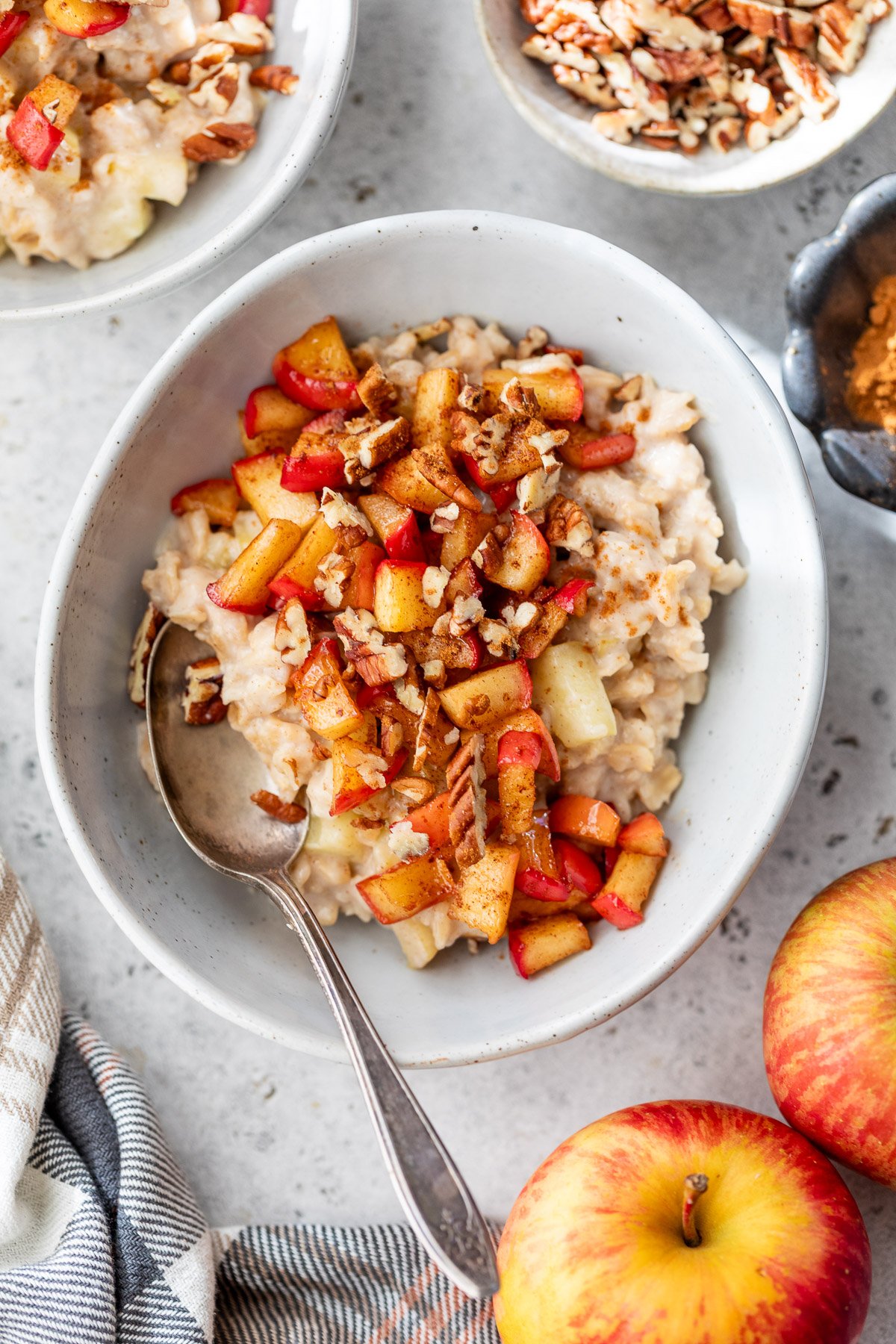 A bowl of apple cinnamon oatmeal topped with chopped apples, pecans, and cinnamon, with a spoon beside it and apples nearby.