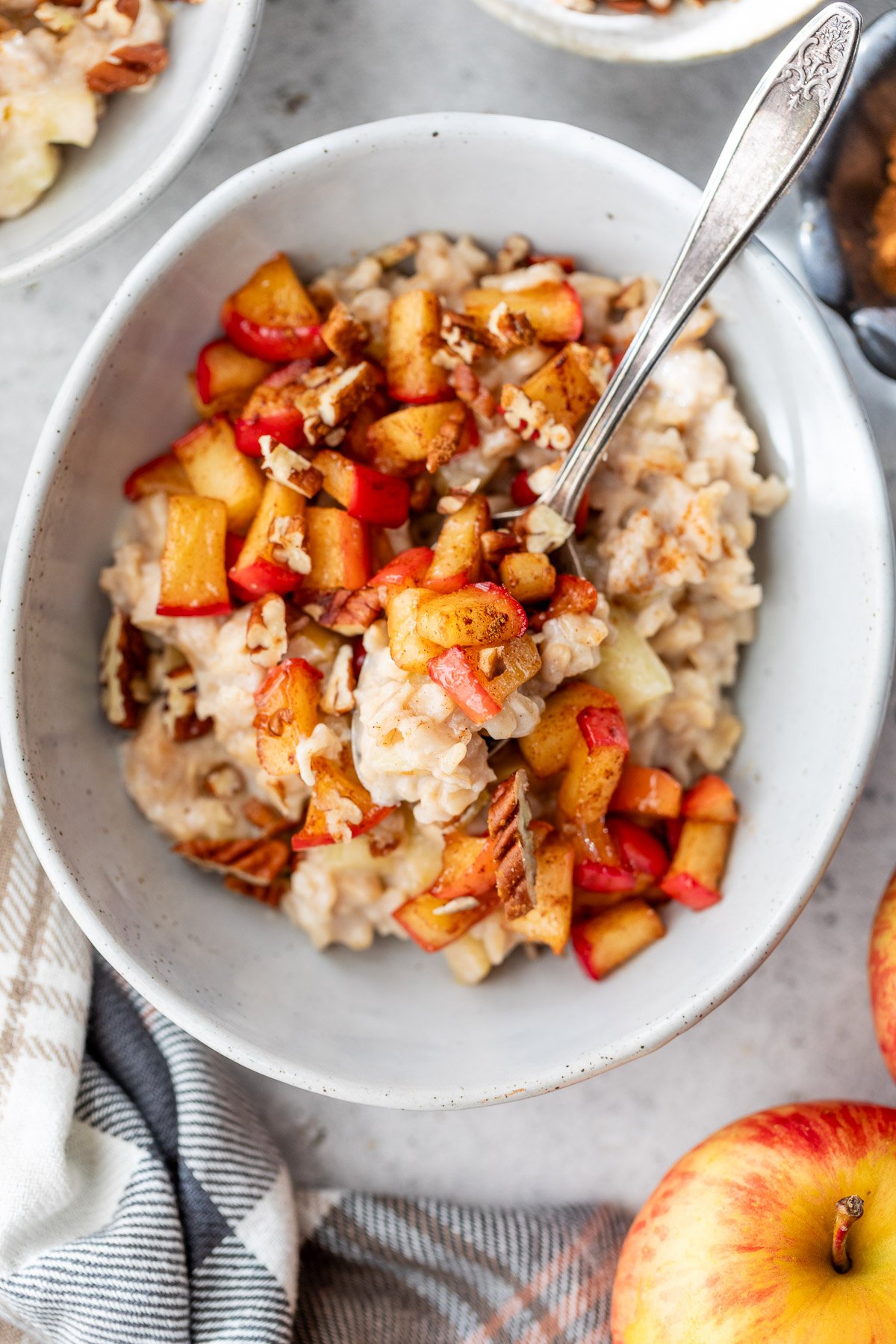Bowl of apple cinnamon oatmeal topped with diced apples, pecans, and cinnamon, with a spoon, on a light table.