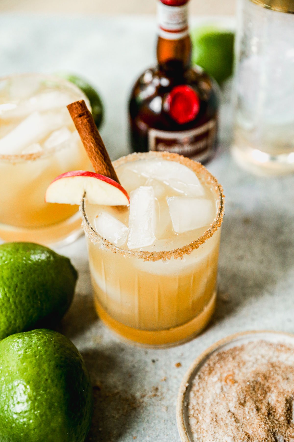 An apple cider margarita with ice, a cinnamon stick, and apple slice garnish in a rimmed glass, surrounded by limes and a liqueur bottle.