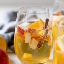 A glass of apple cider sangria with ice, apple and orange slices, and a cinnamon stick, on a light surface.