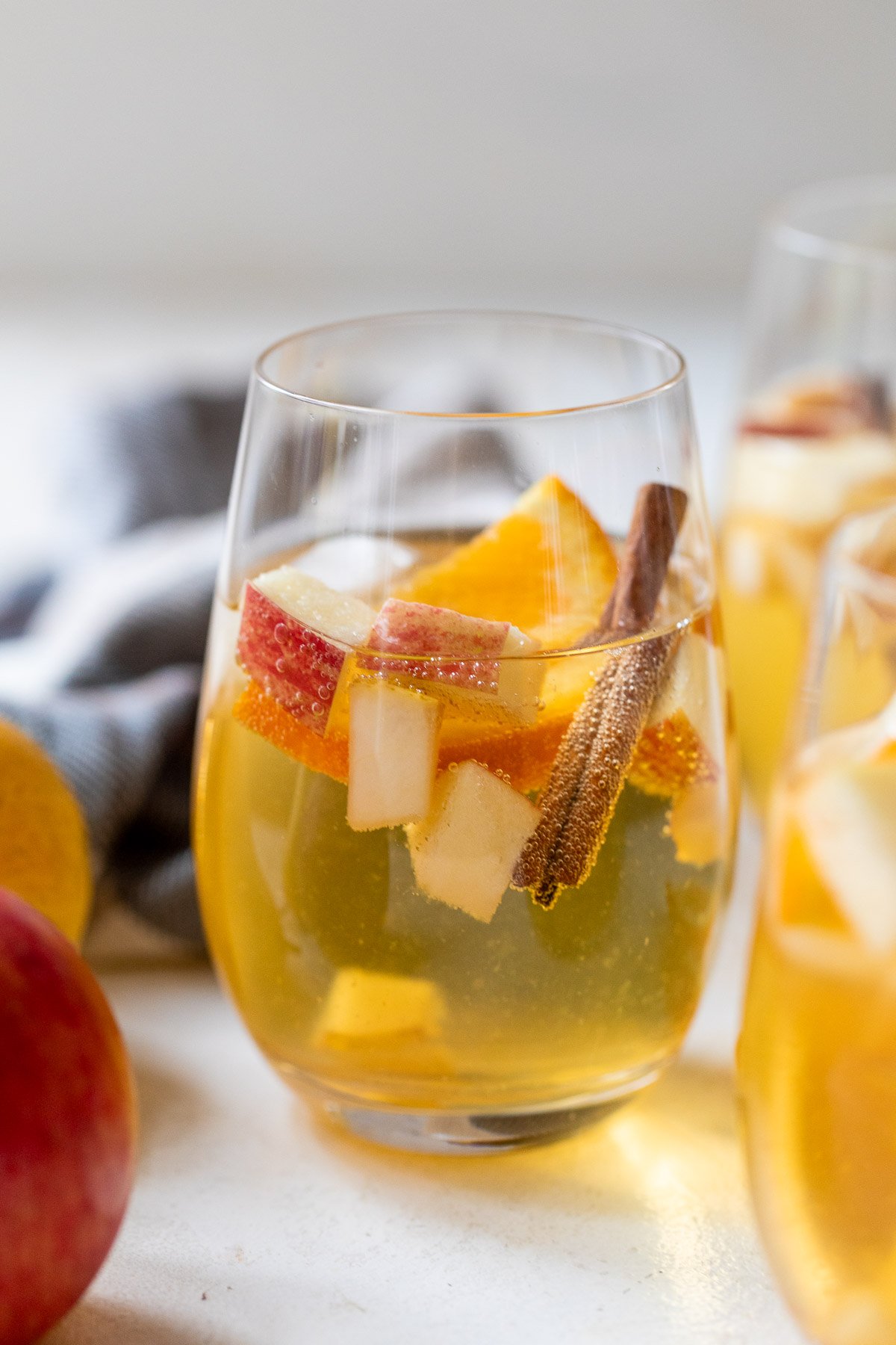 A glass of apple cider sangria with ice, apple and orange slices, and a cinnamon stick, on a light surface.
