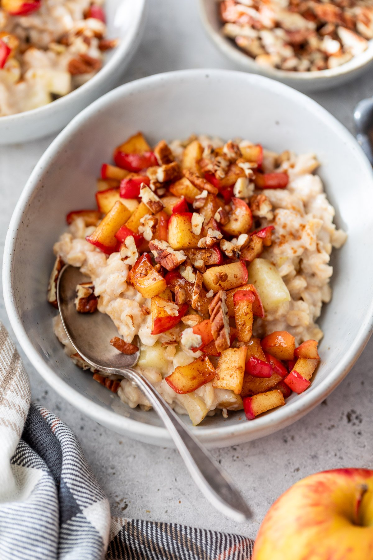 A bowl of apple cinnamon oatmeal topped with diced apples, chopped pecans, and cinnamon, with a spoon resting inside.