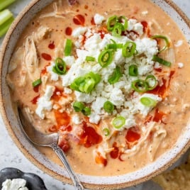 A bowl of creamy Buffalo chicken chili topped with crumbled cheese, chopped green onions, and a drizzle of hot sauce.