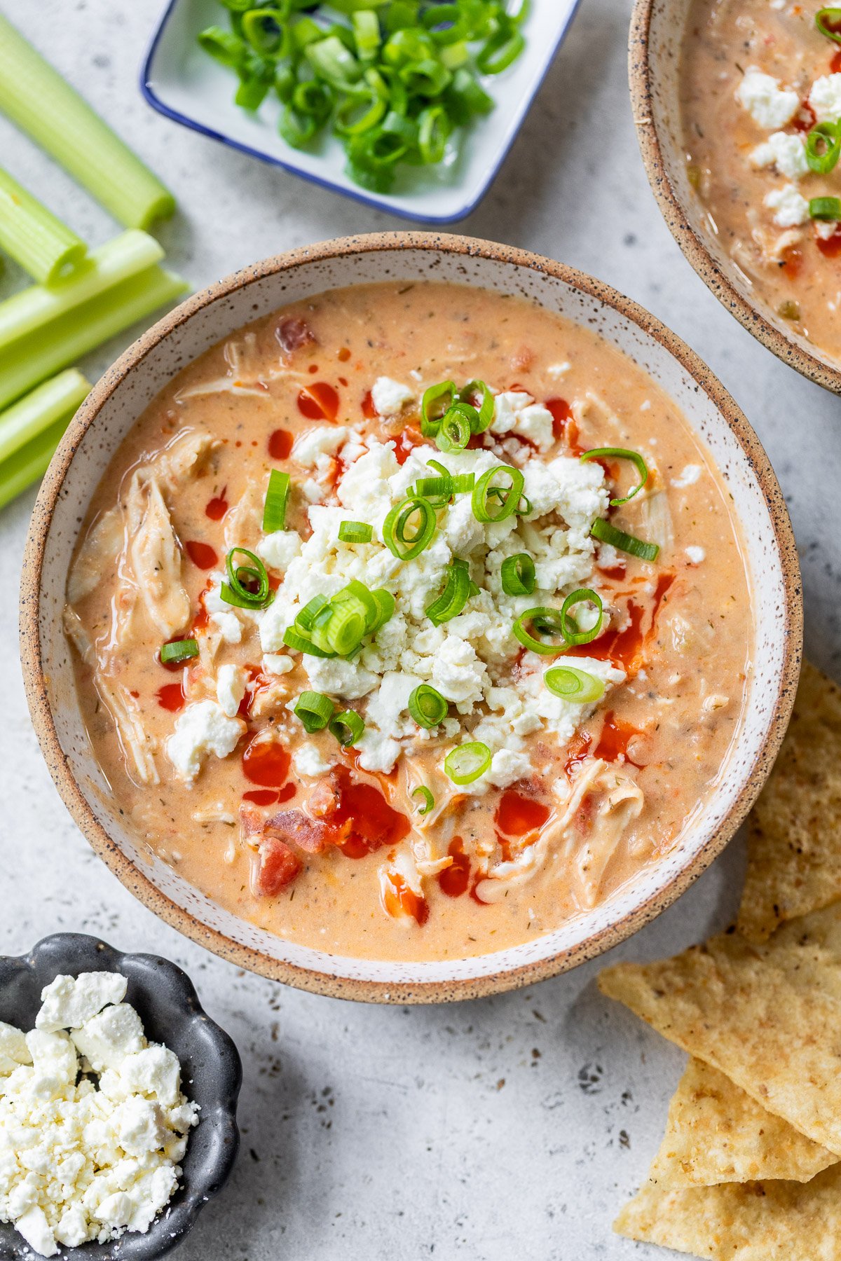 Bowl of creamy Buffalo chicken chili topped with crumbled cheese, hot sauce, and chopped green onions, with chips and veggies nearby.