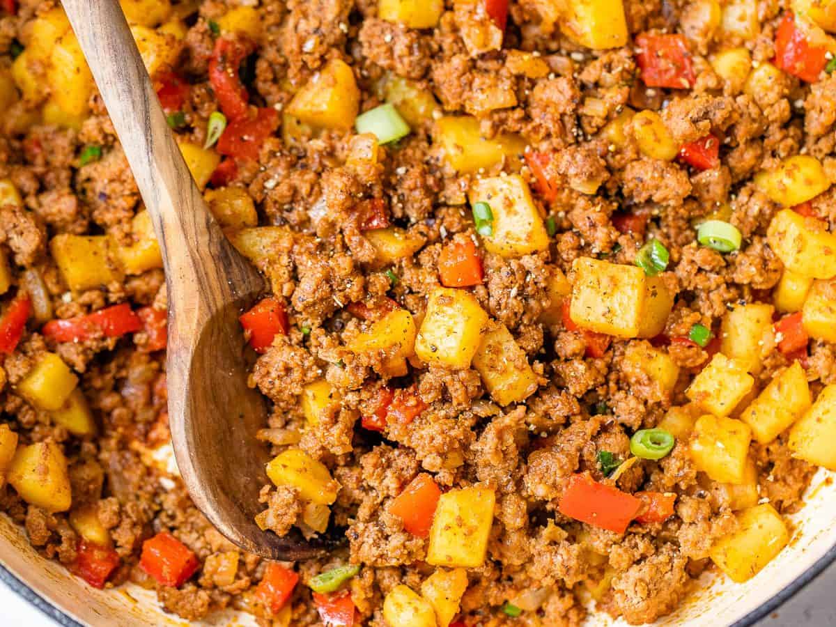 A wooden spoon stirs a skillet of cooked ground beef, diced potatoes, red bell peppers, and green onions.