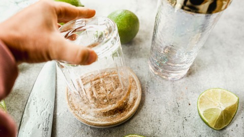 A hand dips a glass into a dish of cinnamon sugar salt with limes and a knife on a countertop nearby.