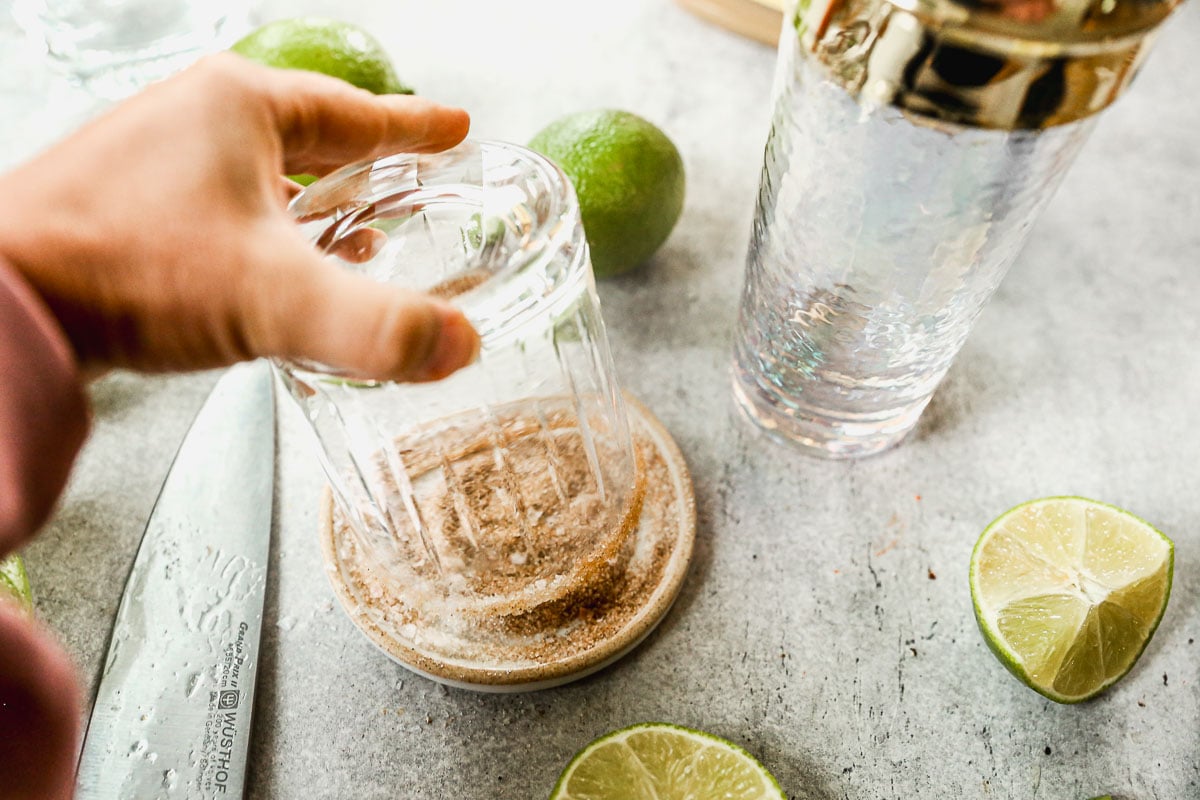 A hand dips a glass into a dish of cinnamon sugar salt with limes and a knife on a countertop nearby.