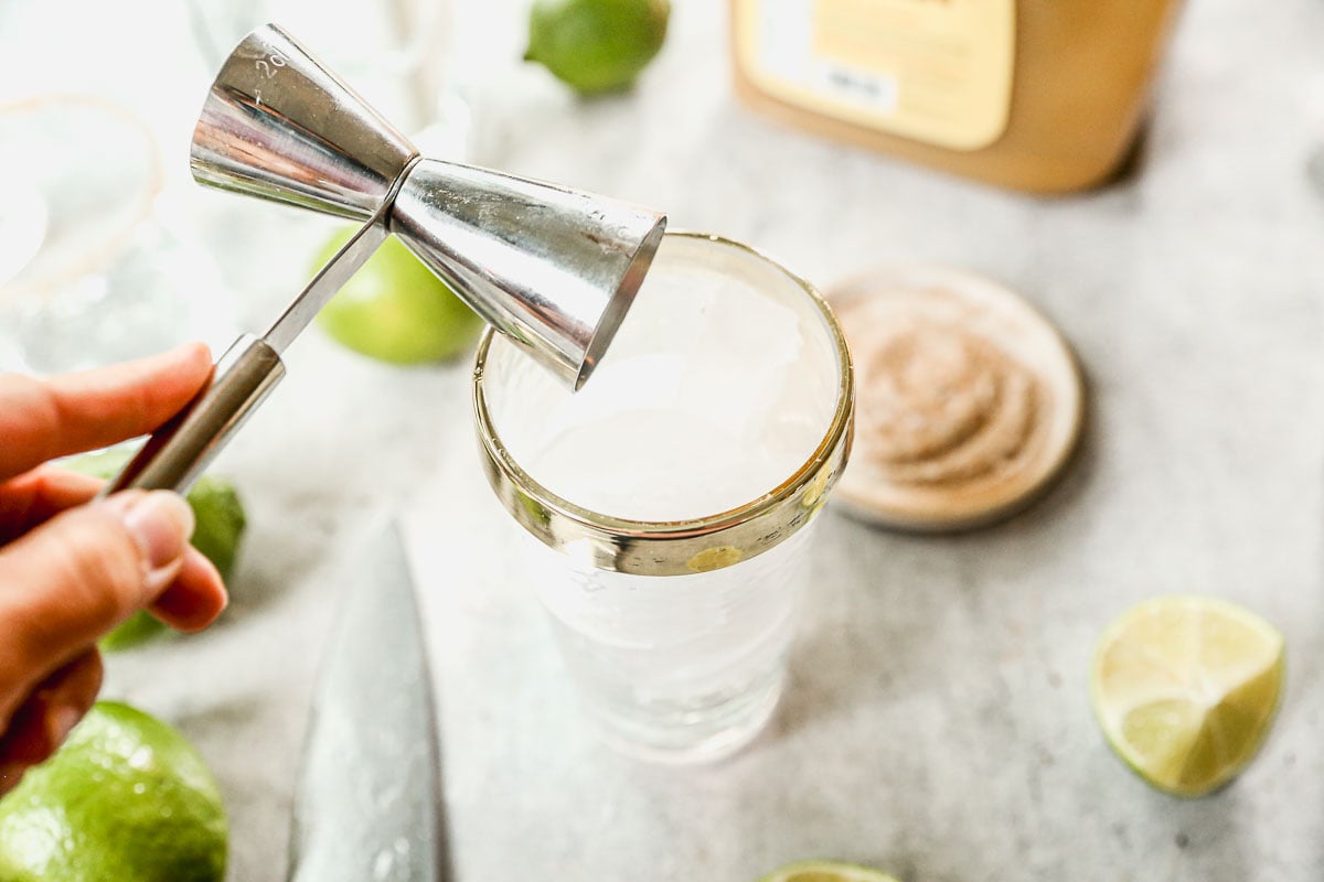 A hand holds a jigger above a glass with a gold rim, surrounded by limes, a knife, and a container.