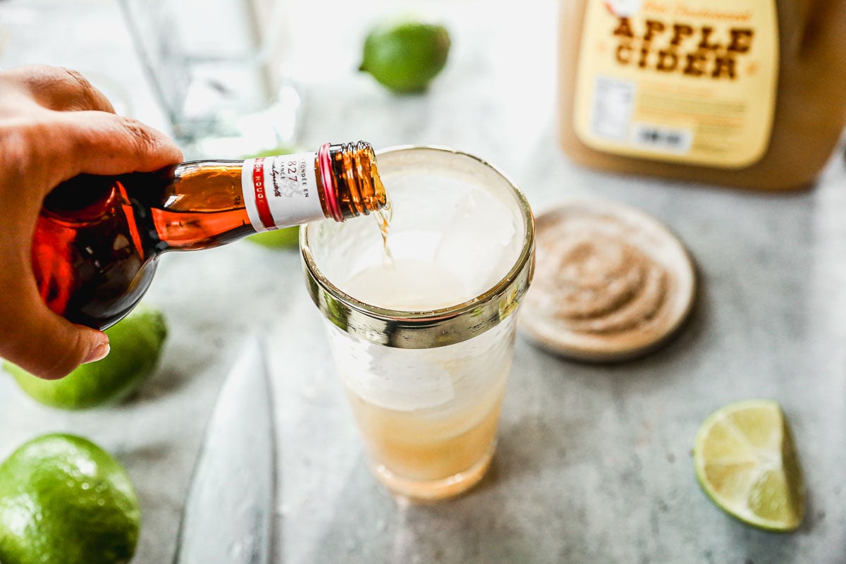 A hand pours liquor into a glass with ice, surrounded by limes, apple cider, and a plate of cinnamon sugar.