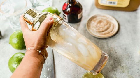 A hand holding a cocktail shaker with ice, surrounded by limes, a bottle, and a sugar-rimmed glass on a countertop.