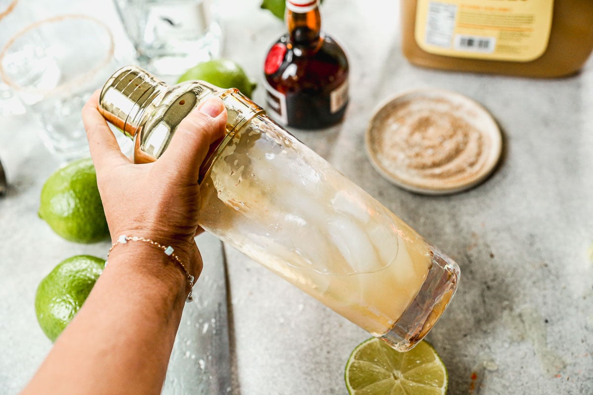 A hand holding a cocktail shaker with ice, surrounded by limes, a bottle, and a sugar-rimmed glass on a countertop.