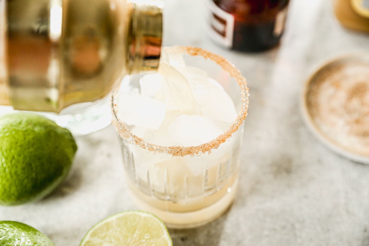 An apple cider margarita is being poured over ice into a glass with a spiced rim, surrounded by fresh lime halves.