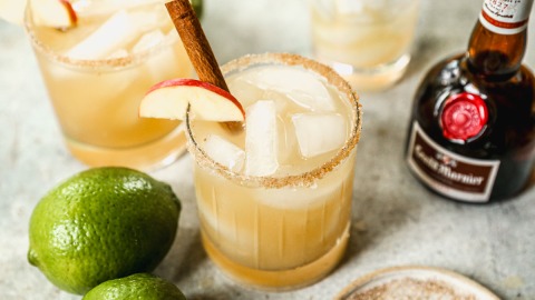 Two glasses of apple cider margarita with cinnamon sticks and apple slices, surrounded by limes and a bottle of Grand Marnier.