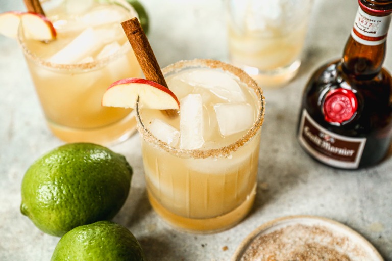 Two glasses of apple cider margarita with cinnamon sticks and apple slices, surrounded by limes and a bottle of Grand Marnier.