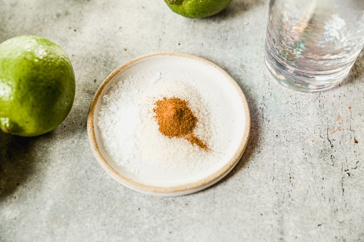 A small plate with salt, sugar, and cinnamon, next to whole limes and a glass of water on a gray surface.
