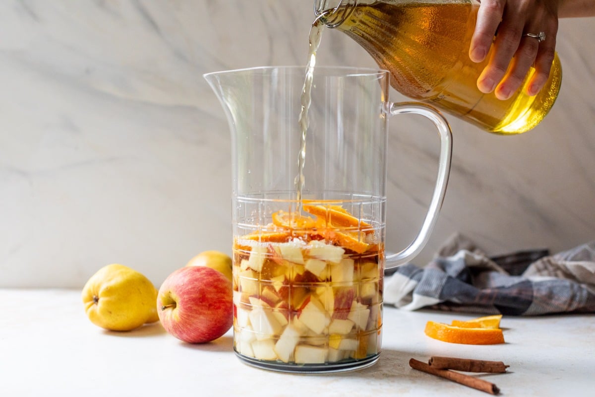 A hand pours wine into a pitcher with chopped apples, oranges, and cinnamon sticks. Apples and cinnamon are nearby.