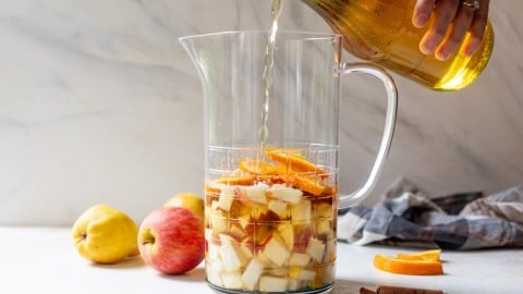 A hand pours wine into a pitcher with chopped apples, oranges, and cinnamon sticks. Apples and cinnamon are nearby.