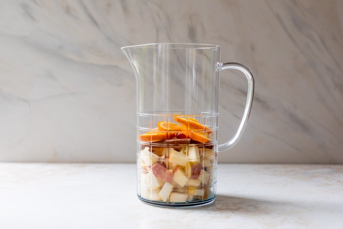 Clear pitcher with chopped apples, orange slices, and cranberries layered inside, placed on a light countertop.