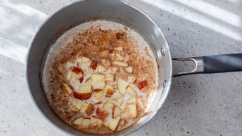 A saucepan with chopped apples, cinnamon, and oatmeal simmering on a light-colored countertop.
