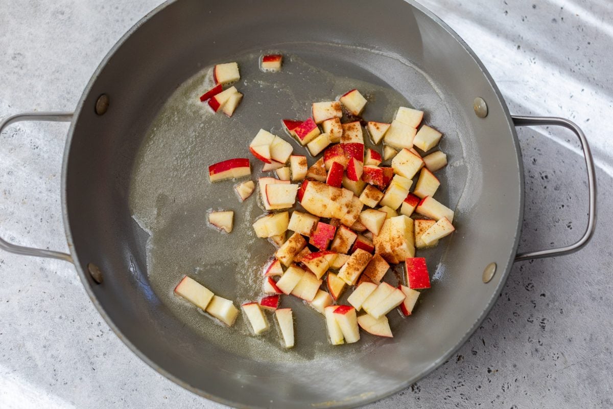 Chopped red apples with cinnamon cooking in a gray skillet on a light countertop.